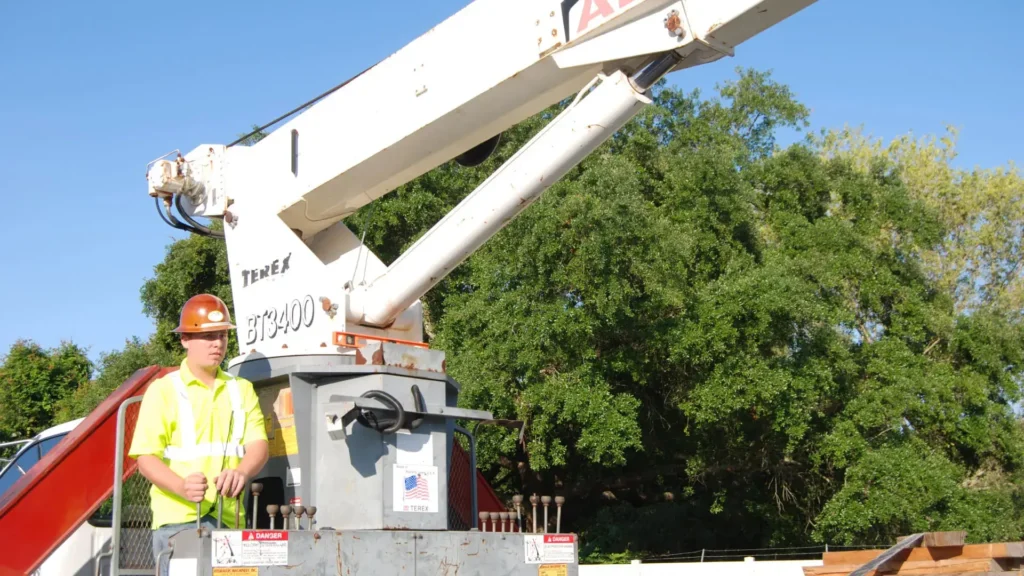 A worker in a bright yellow safety vest and hard hat operates a white boom truck crane.