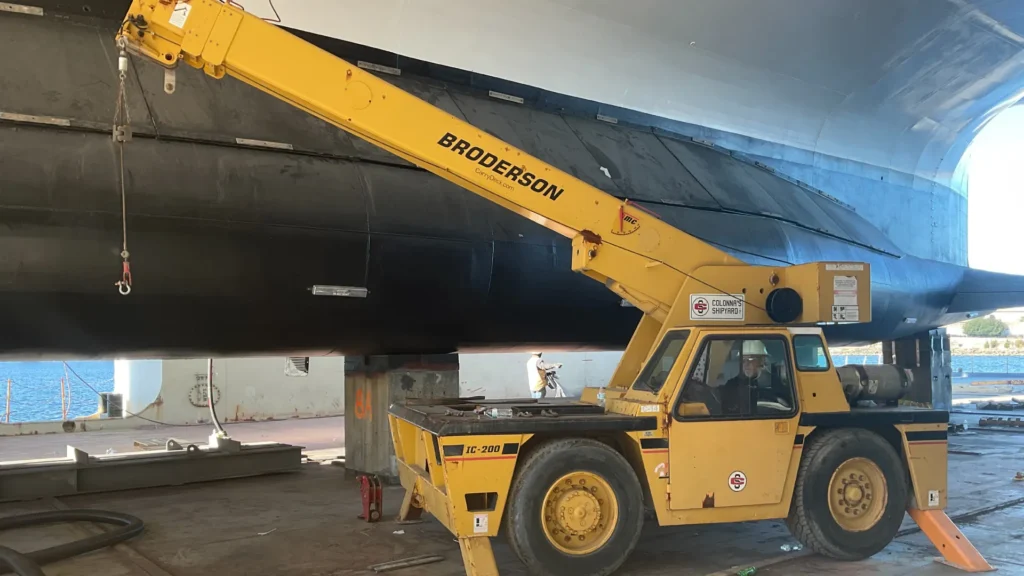 A yellow industrial crane labeled is positioned on a ship's deck, lifting or stabilizing a large black structure. The setting is adjacent to water, under a large archway, giving a sense of carry deck crane training.