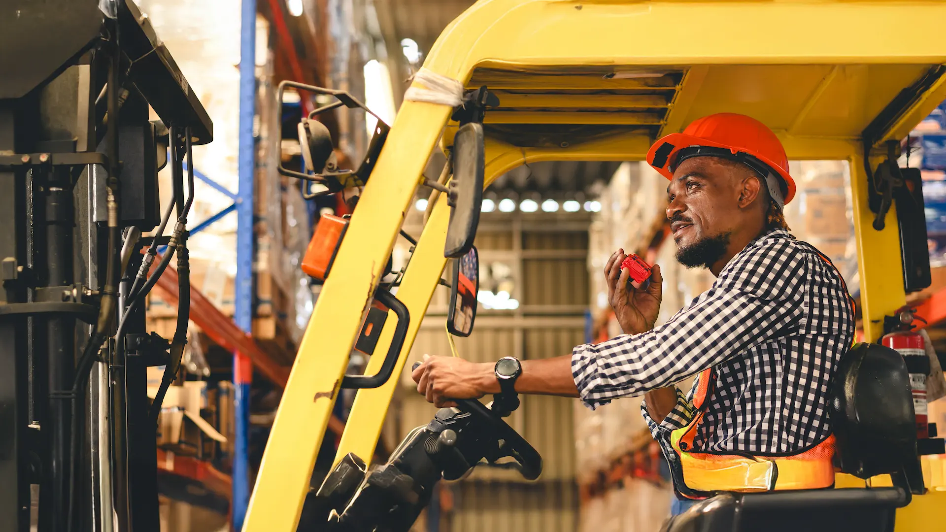 A man in an orange hard hat operates a yellow forklift in a warehouse. He holds a radio. Represents Forklift Operator Training