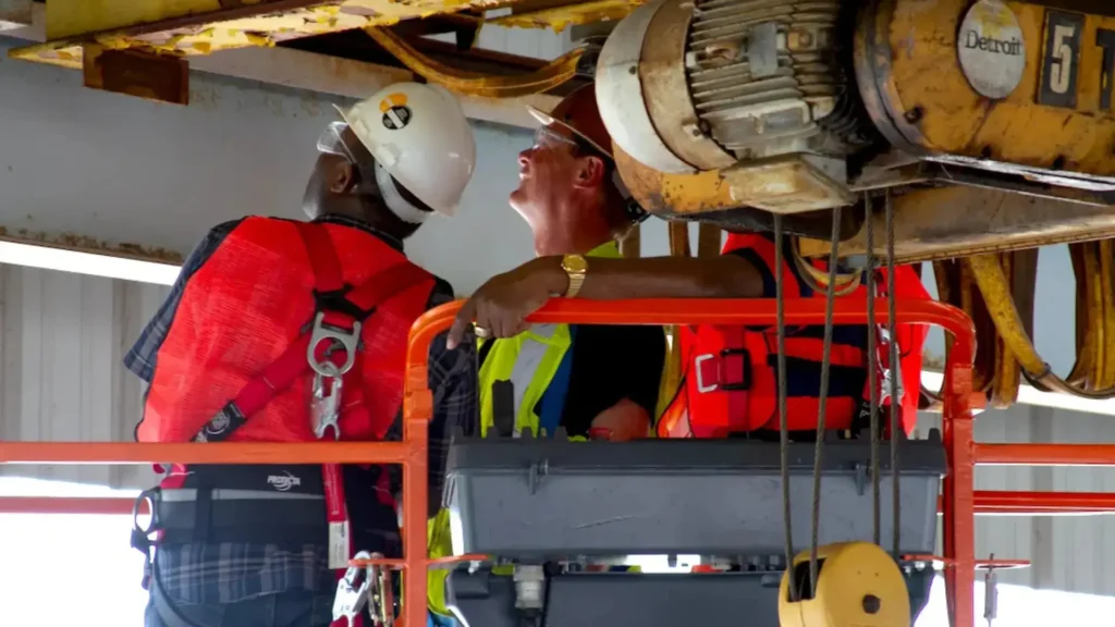 Two construction workers in safety gear, including helmets and red vests, inspect the overhead crane closely. The setting is industrial, and safety-oriented.