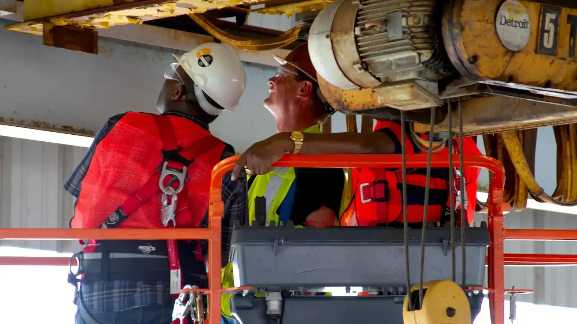 Two construction workers in safety gear, including helmets and red vests, inspect the overhead crane closely. The setting is industrial, and safety-oriented.