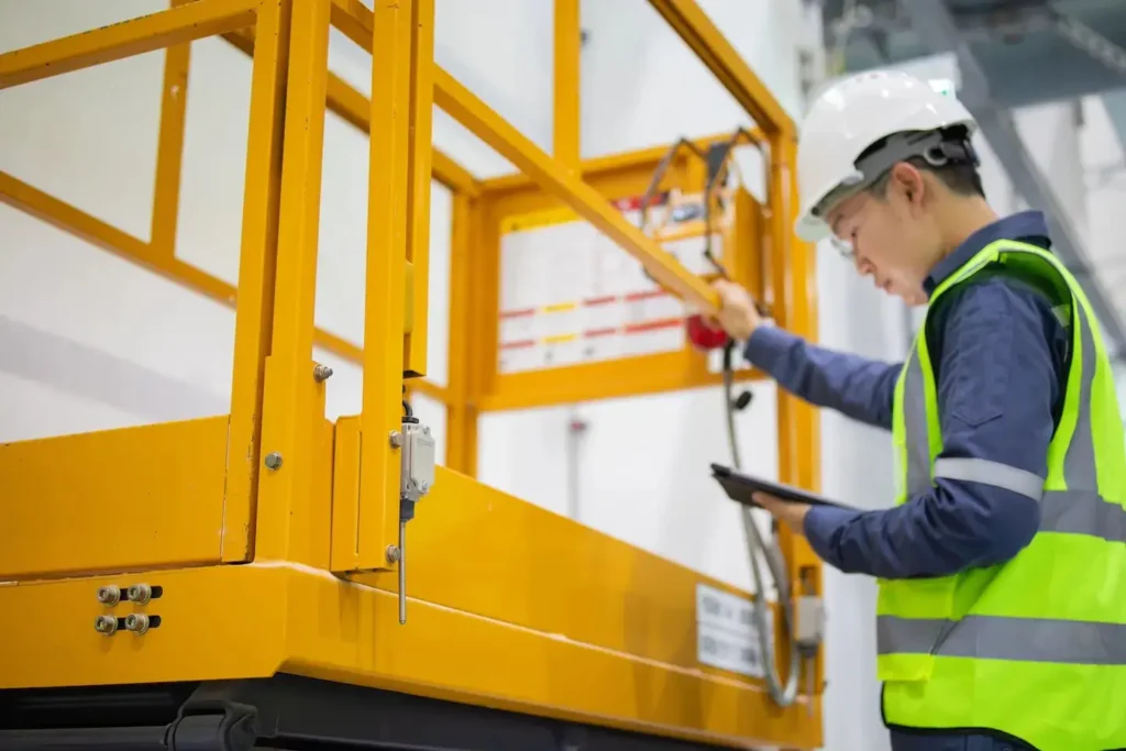 A worker in safety gear, including a hard hat and reflective vest, inspects a MEWP aerial lift with a tablet.