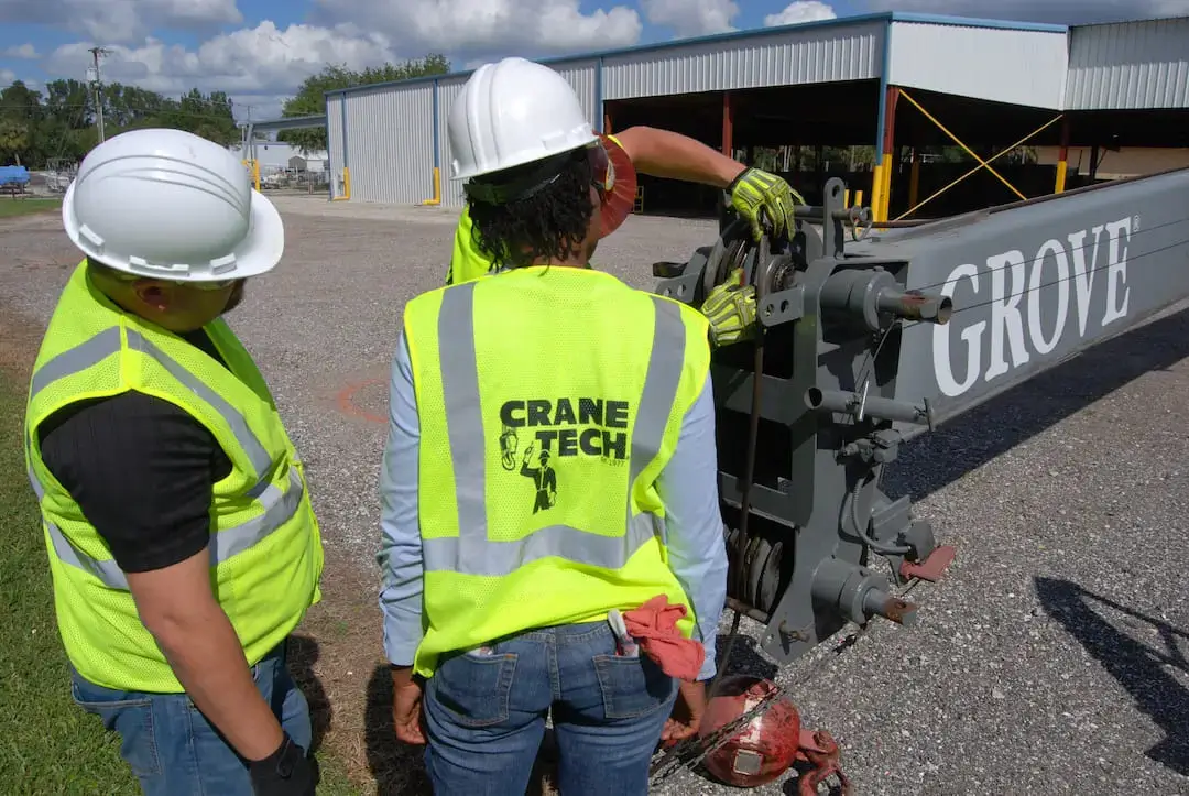 Three construction workers in hard hats and neon vests labeled "CRANE TECH" inspect a mobile crane outdoors. One points at the mechanism, discussing safety.