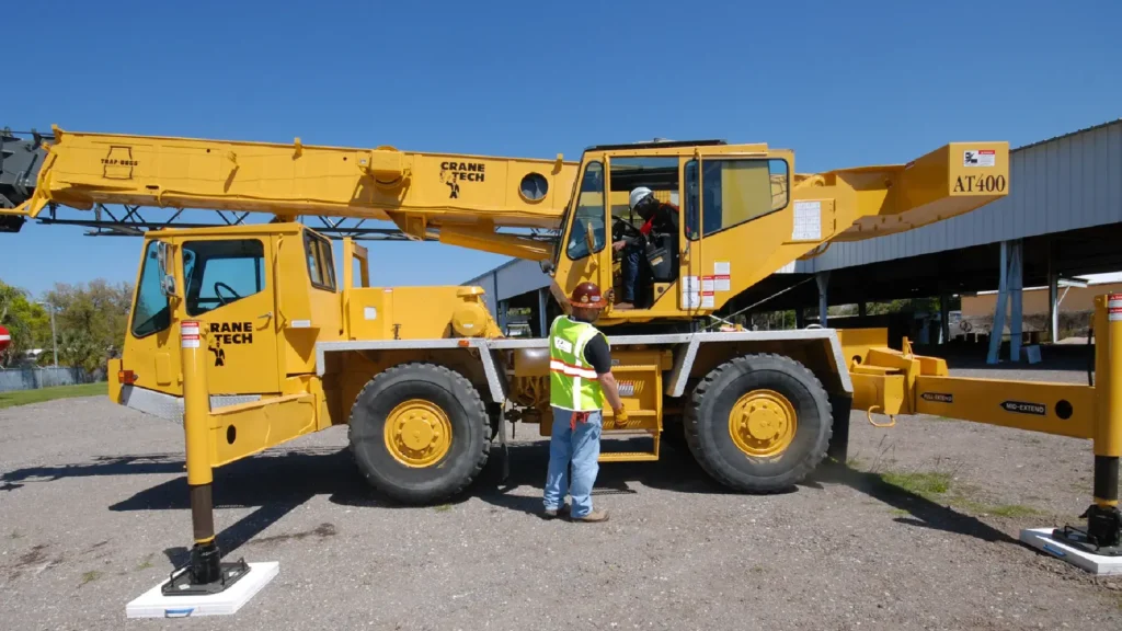 A yellow mobile crane with "Crane Tech" branding is parked on a gravel lot. A man in a safety vest and helmet stands beside it.