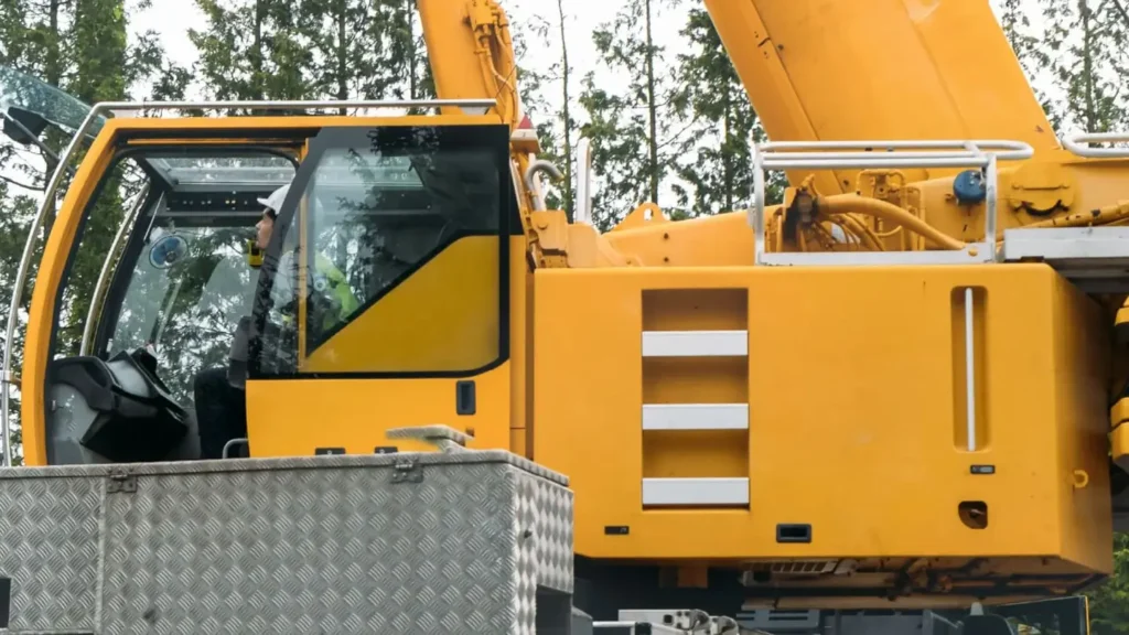 A yellow construction mobile crane with a person inside the cab operates against a backdrop of green trees. The scene conveys the mobile crane operator training.
