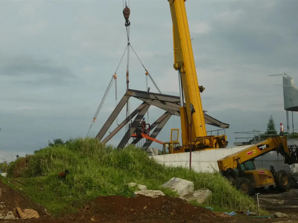 A yellow crane lifts metal beams at a construction site on a cloudy day. Nearby, a worker on a lift oversees the operation, surrounded by grass and dirt.