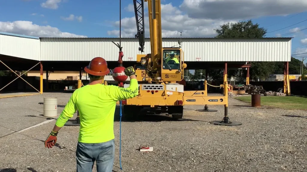 A construction worker in a neon shirt and hard hat guides a yellow crane with a hand signal on a gravel lot, under a partly cloudy sky.