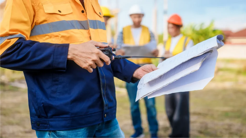 A construction worker in an orange and blue jacket holds a two-way radio and blueprint. Blurred coworkers in safety gear are in the background.