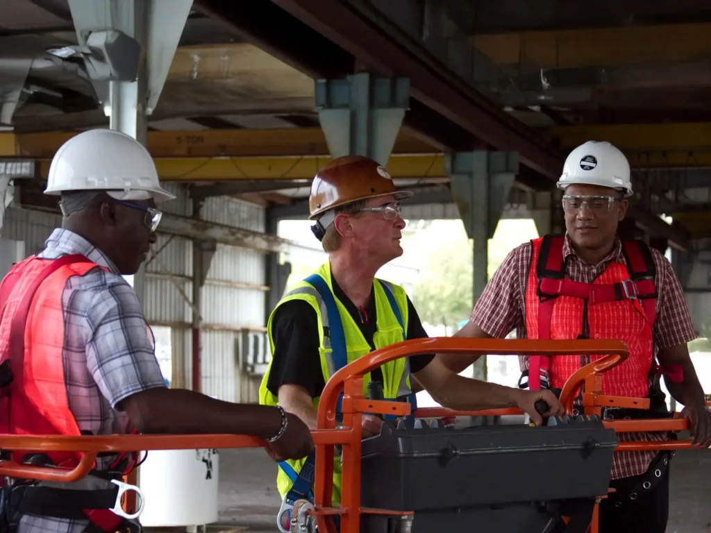 Three construction workers in safety gear, including helmets and vests, perform MEWP Aerial Lift Operator Training inside an industrial building.