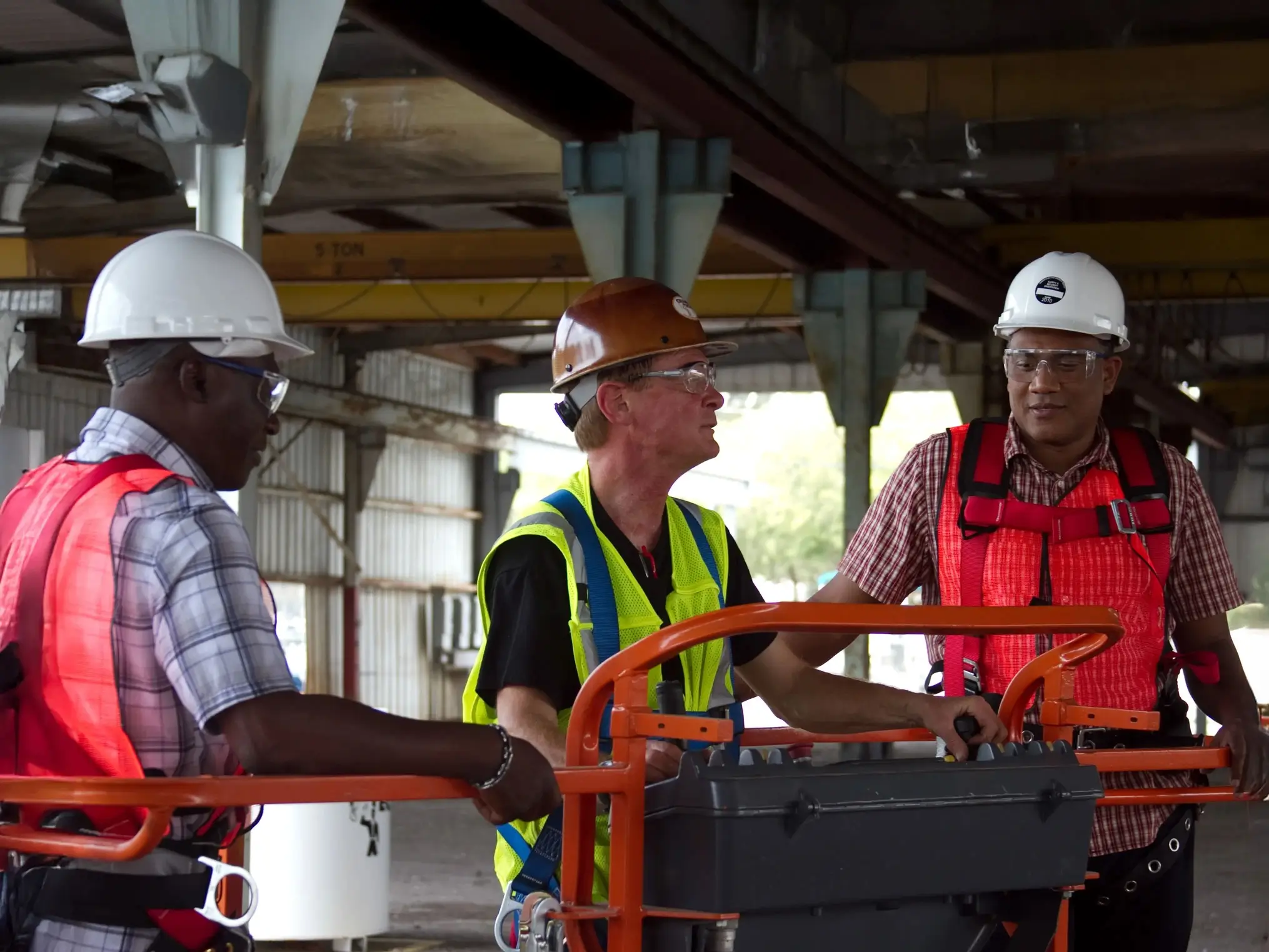 Three construction workers in safety gear, including helmets and vests, perform MEWP Aerial Lift Operator Training inside an industrial building.