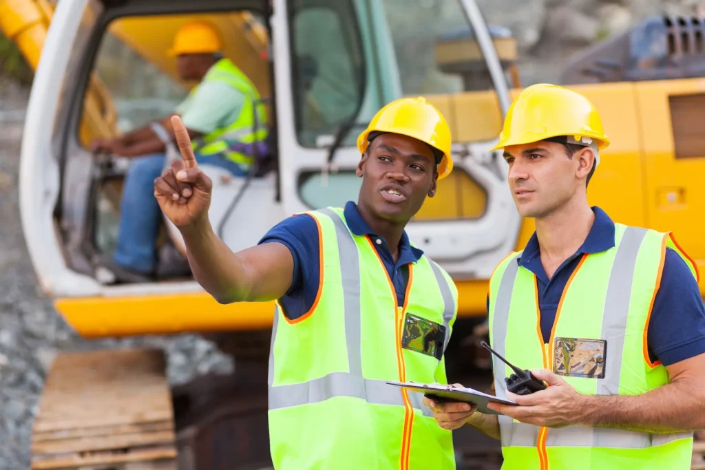 Two construction workers in hard hats and reflective vests discuss plans beside yellow machinery. One points forward, indicating direction. Representes the OSHA 10 Construction Industry Training.
