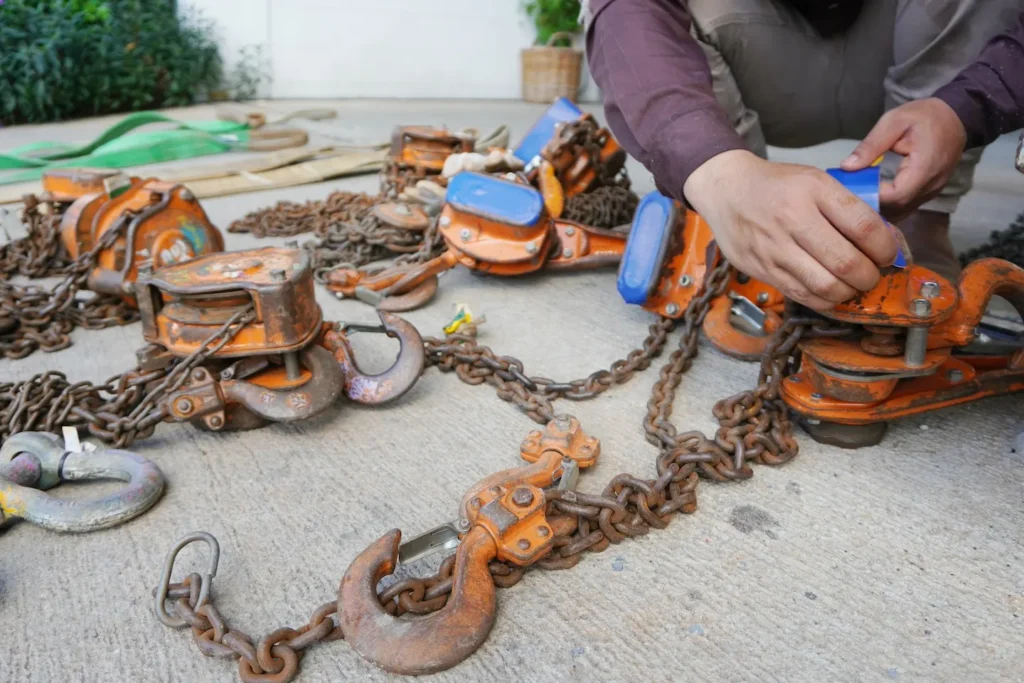 Person squatting on concrete, handling rusty orange and blue chain hoists and hooks. The scene conveys an industrial, hands-on work environment. Rigging Gear Sling Inspector Training