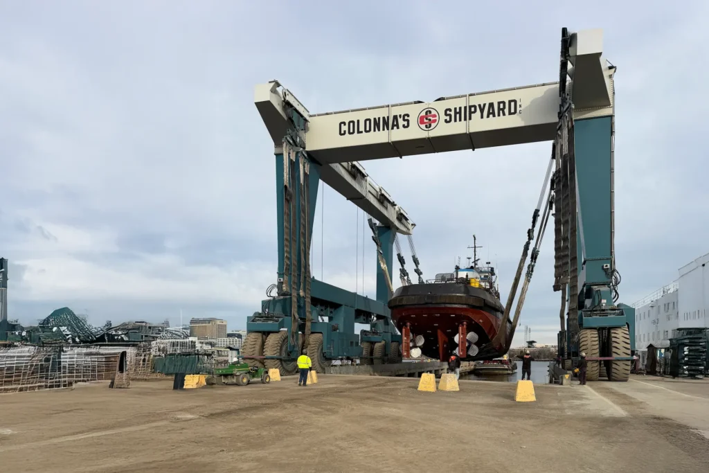 A massive ship hoist at Colonna’s Shipyard lifts a boat. Workers in reflective jackets stand nearby. Portrays the Straddle Crane Operator Training.