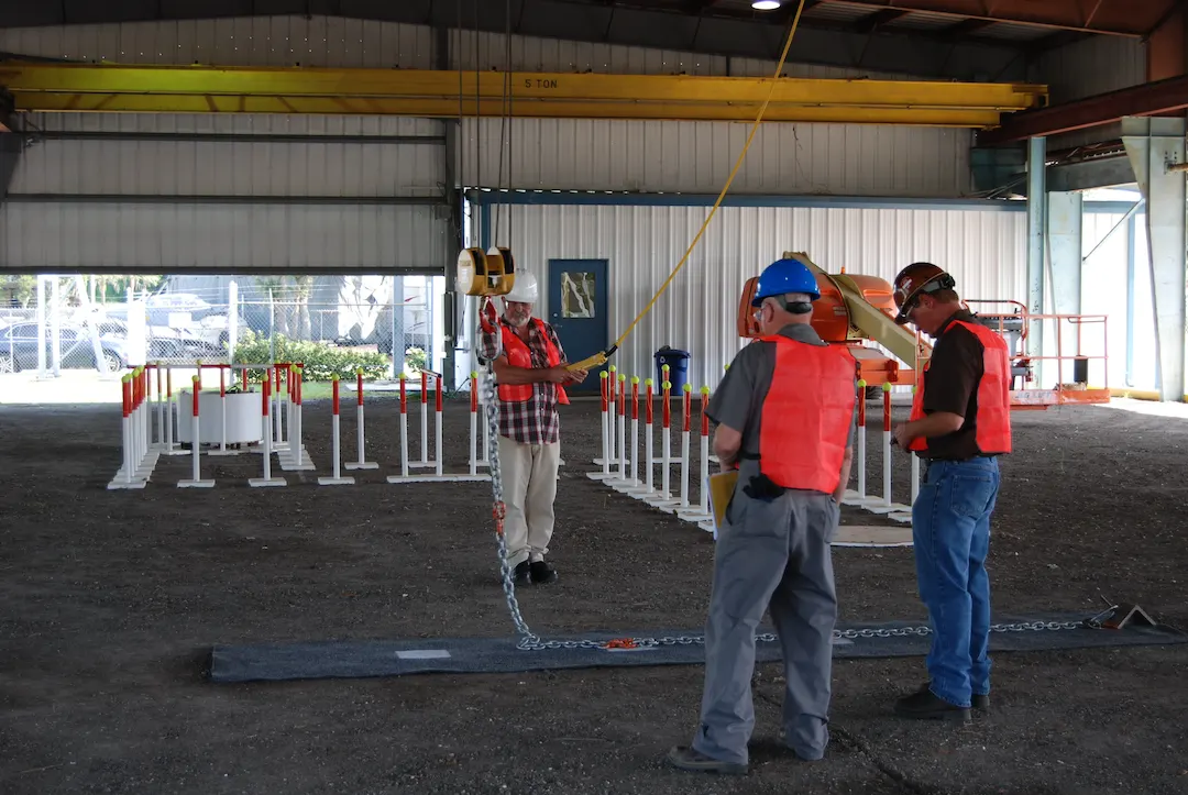 Three construction workers in safety vests and helmets assess a suspended steel beam in a warehouse during the Overhead Gantry Crane Operator Training.