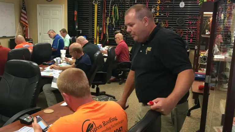 A man in a black Crane Tech polo conducts a small workshop in a conference room. Participants sit at a table, focused on notes and devices. The room has a casual, collaborative atmosphere.