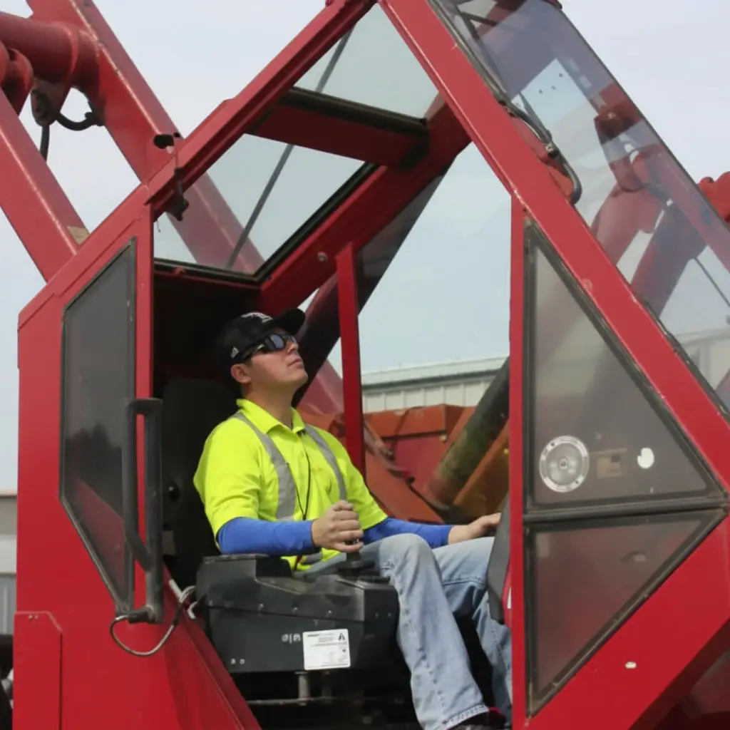 A construction worker in a safety shirt operates a red crane from inside the cab.